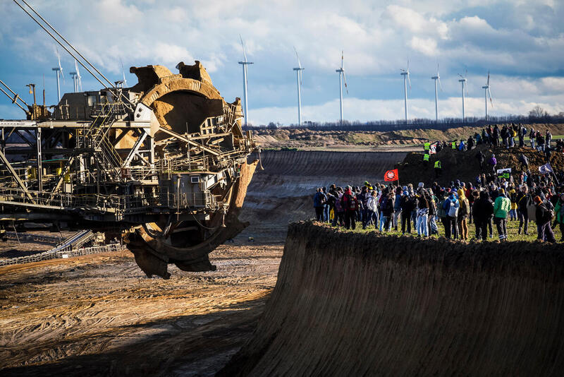 Zu sehen ist ein riesiger Kohlebagger und eine Menschenmeng, die am Rand der Kohlegrube bei Lützerath stehen und gegen das abbaggern protestieren.