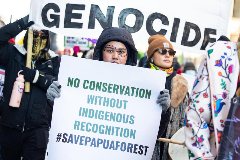 Eine indigene Frau steht in einer Menschenmenge und hält ein Plakat mit der Aufschrift: 